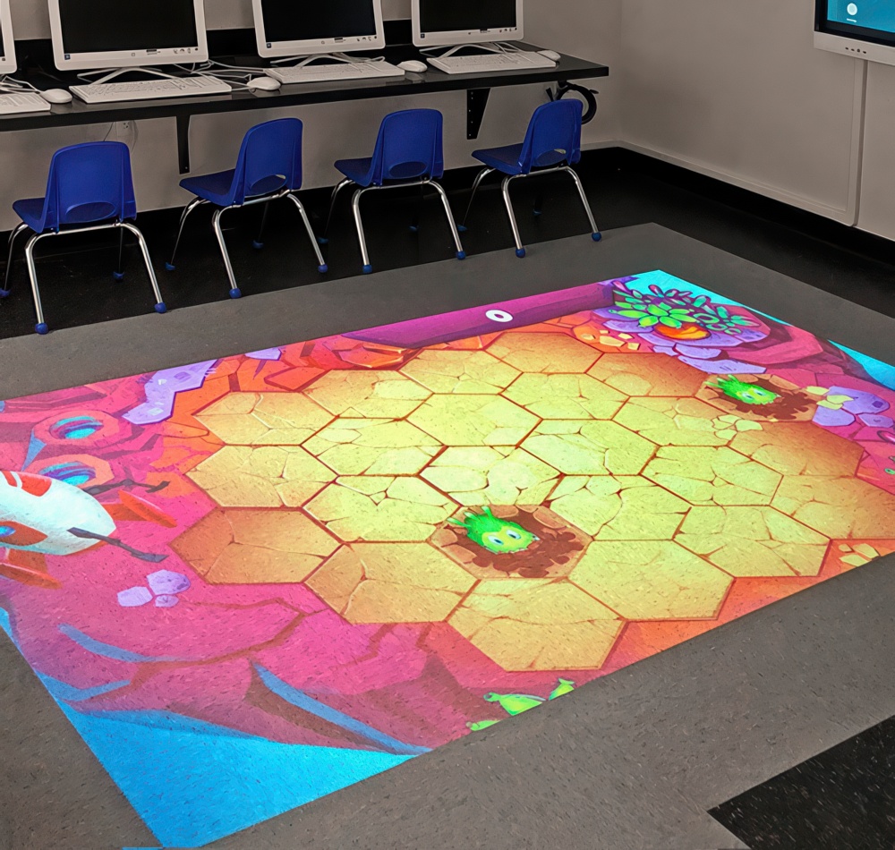 Interactive floor projection displaying a colorful game scene with hexagonal tiles, set up in a classroom with blue chairs.