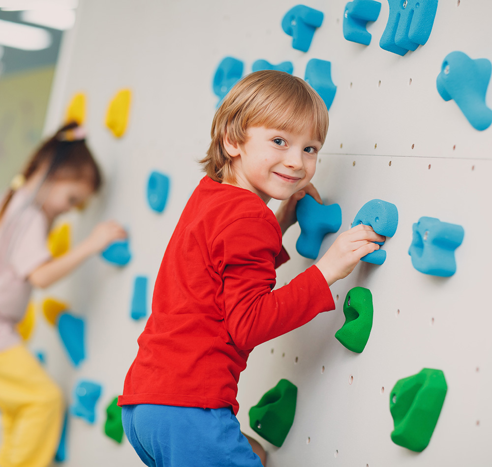A smiling boy is climbing a colorful indoor rock wall, showing excitement and determination as he navigates the holds.