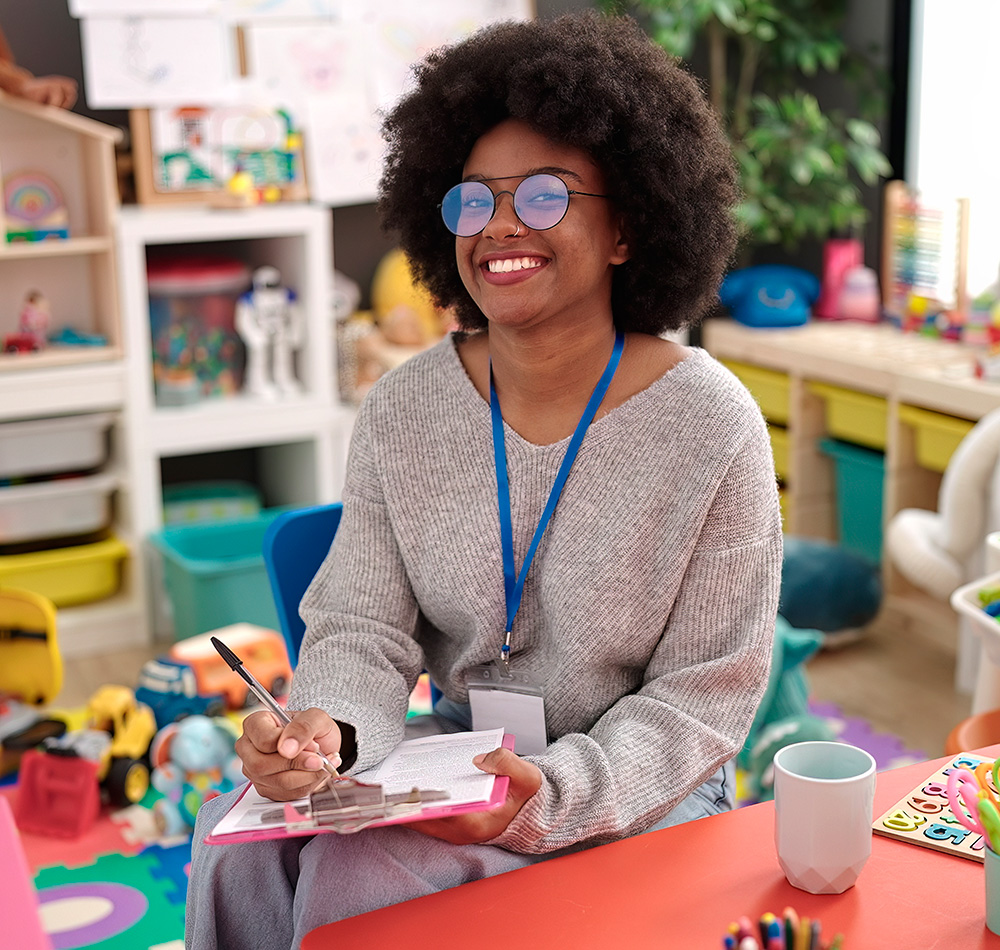 A smiling teacher with glasses is sitting in a colorful classroom, holding a clipboard and pen.