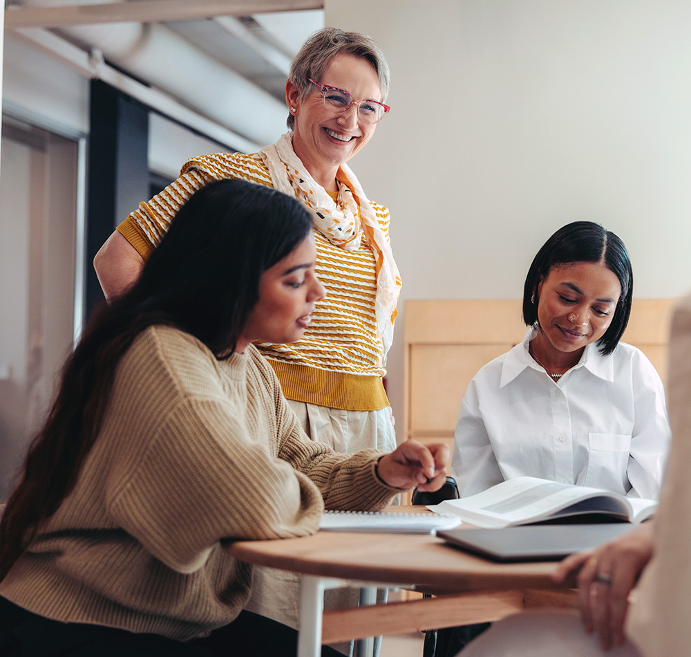 An older teacher is standing and smiling while observing two younger teacher who are focused on their studies.